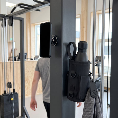 Gympaketet i bruk: mobilen i ögonhöjd på den magnetiska mobilhållaren, flaskan i den magnetiska gymväskan och handduken på handdukskroken vid en kabelmaskin
