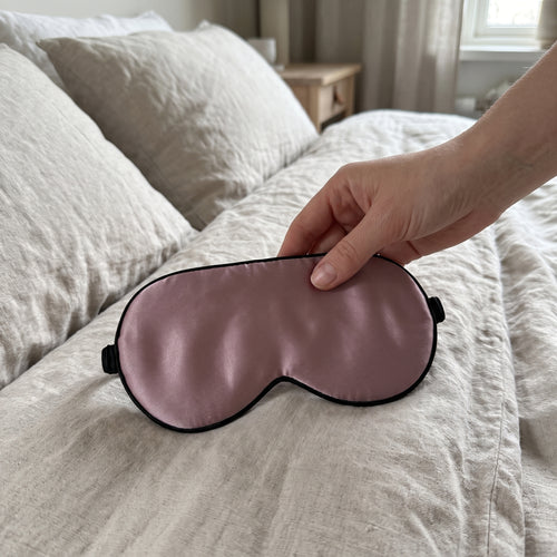 Sovmask i mullbärssilke i dämpad gammelrosa med svart kant och smalt elastiskt band, liggande på en hand