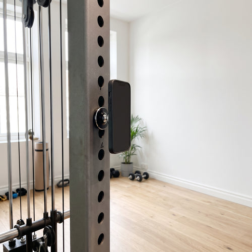 Den svarta magnetiska mobilhållaren på gymställningens stålram med mobilen fäst, i ett ljust svenskt gym