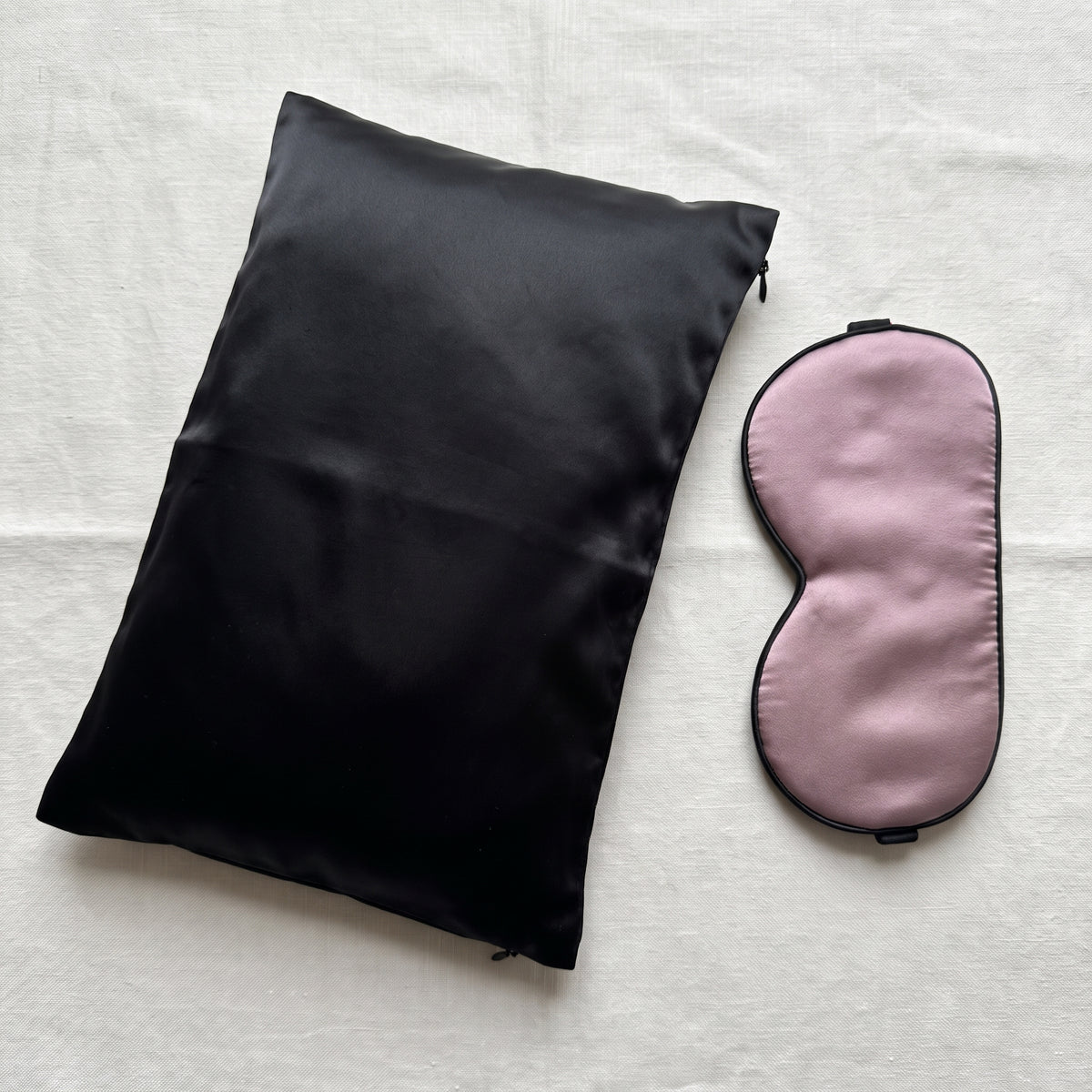Sömnset i mullbärssilke: svart silkesörngott med dragkedja och dammig mauve sovmask med svart kant, utlagda på ljus bakgrund